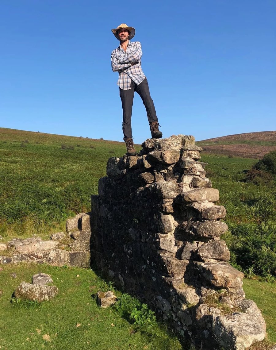 Tom Cox in a wide-brimmed hat, standing on top of a ruined stone wall on open moorland